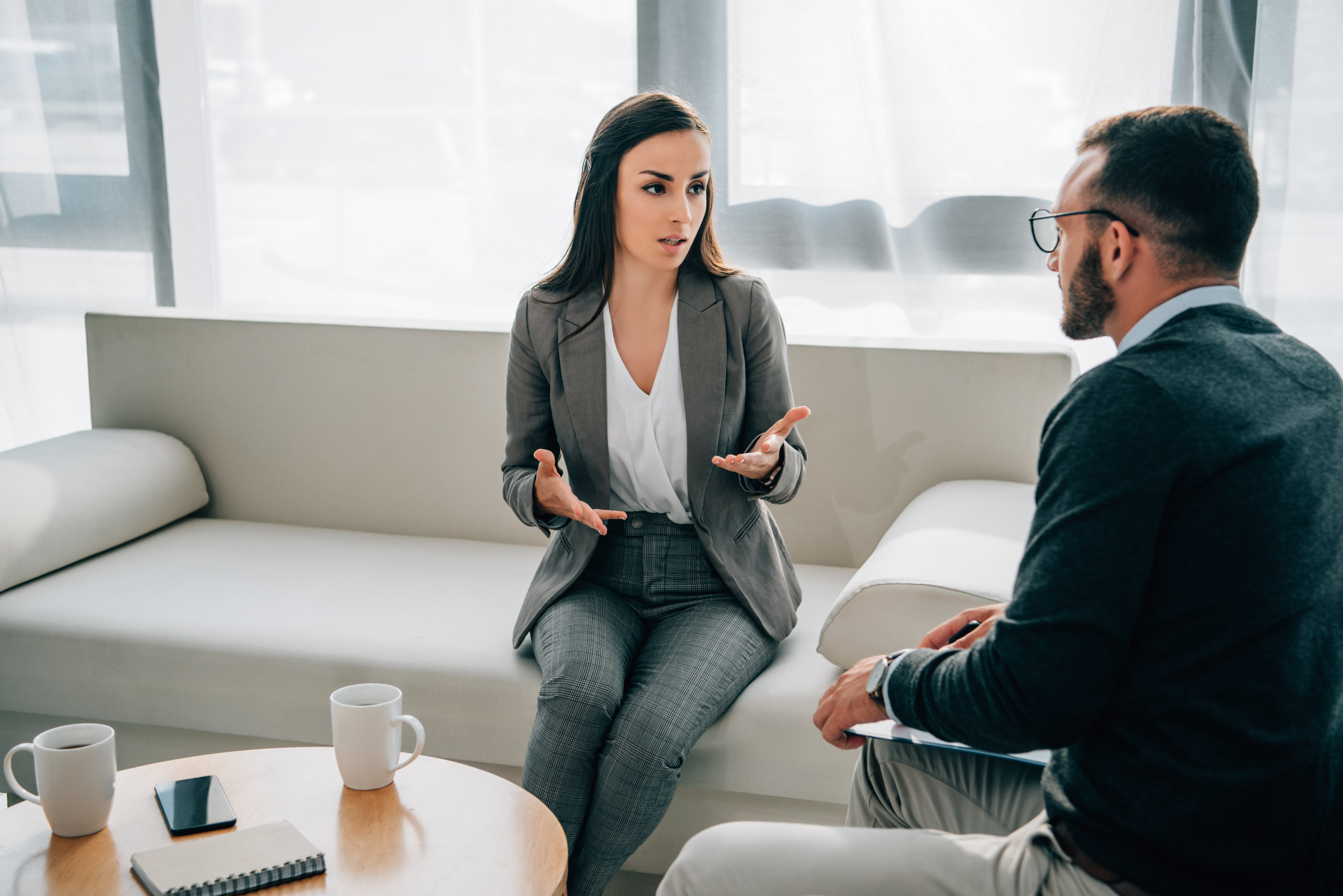 Woman in a business suit sitting on a couch talking to a man sitting down and listening.