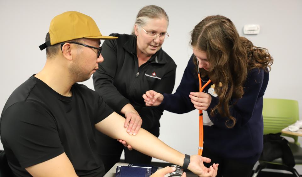 A woman checks a man's pulse, while a teacher watches.