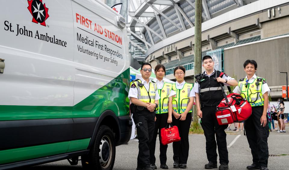 Group of St. John Ambulance volunteers in high-visibility yellow vests standing beside a white ambulance-style van at an outdoor event, with a large building in the background.