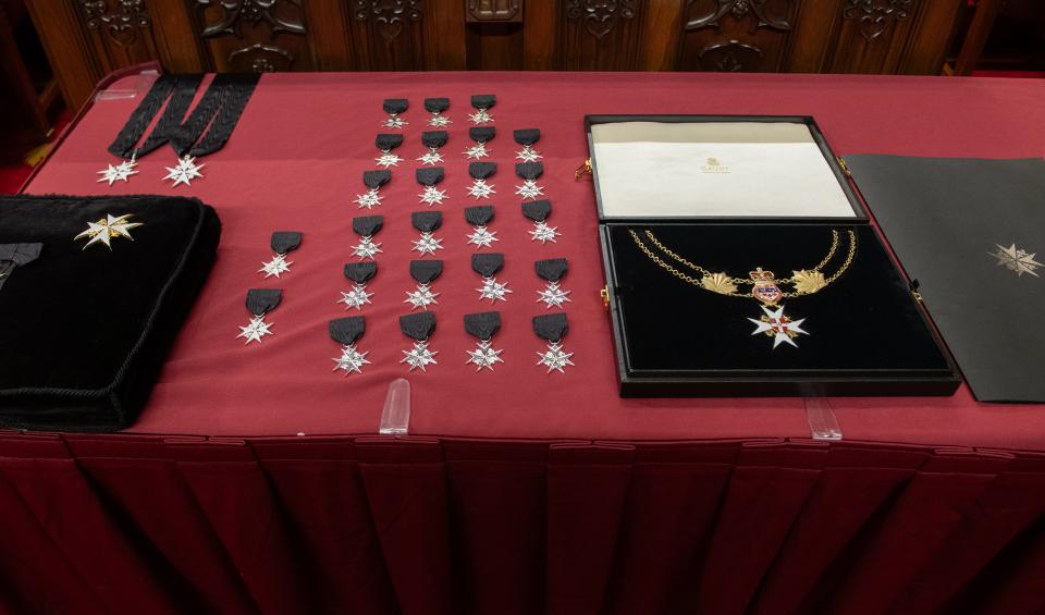 Table covered in a red cloth displaying Order of St John insignia: rows of silver eight-pointed cross medals on black ribbons, two neck badges on black sashes, a black velvet cushion holding gold and white crosses, and an open case with a gold collar chain and cross.