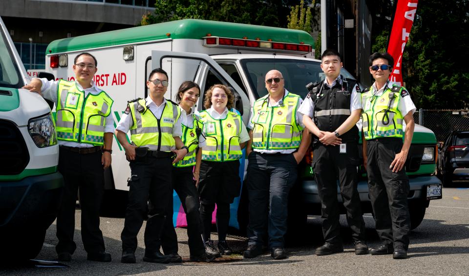 Seven St. John Ambulance first aid volunteers in high-visibility yellow vests and uniforms pose together in front of a white and green St. John Ambulance first aid vehicle at an outdoor event, with a red "First Aid" banner flag behind them.