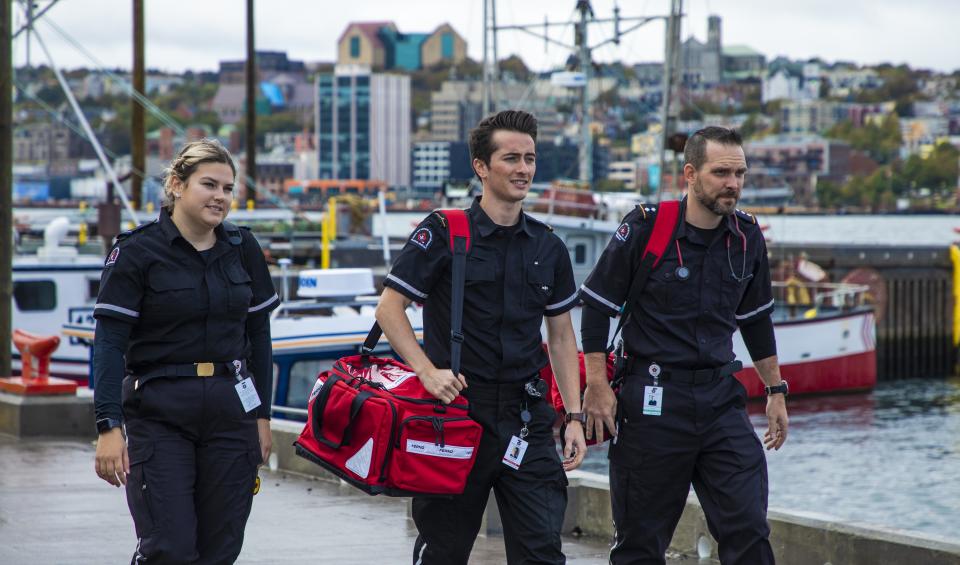 Three St. John Ambulance medical first responders in dark uniforms walk along a harbour dock, one carrying a red medical bag, with fishing boats and a hillside city in the background.