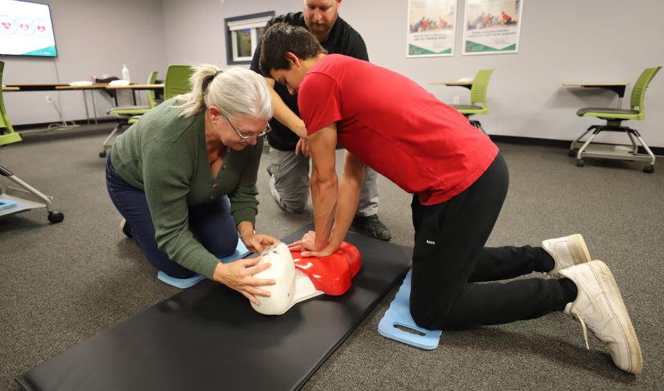 Two students practise CPR on a manikin in a classroom while an instructor guides them.
