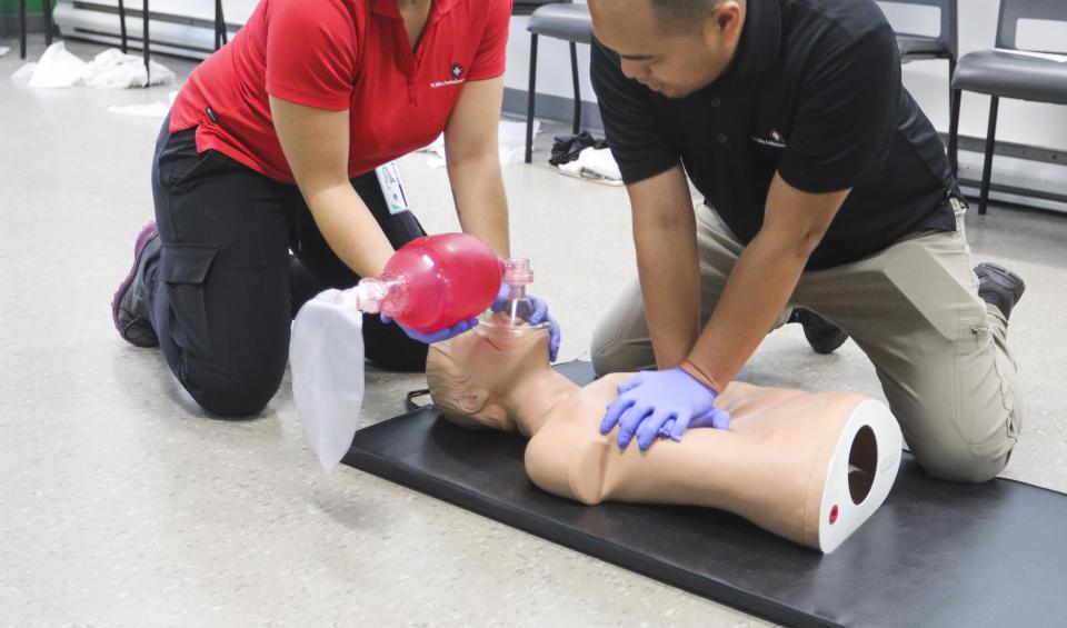Two St. John Ambulance instructors kneel on a mat practising two-rescuer CPR on a training manikin: one gives chest compressions while the other holds a bag-valve mask over the manikin's face. Both wear purple gloves.
