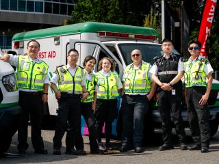 Seven St. John Ambulance first aid volunteers in high-visibility yellow vests and uniforms pose together in front of a white and green St. John Ambulance first aid vehicle at an outdoor event, with a red "First Aid" banner flag behind them.
