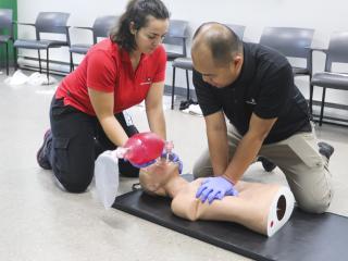 Two St. John Ambulance instructors kneel on a mat practising two-rescuer CPR on a training manikin: one gives chest compressions while the other holds a bag-valve mask over the manikin's face. Both wear purple gloves.