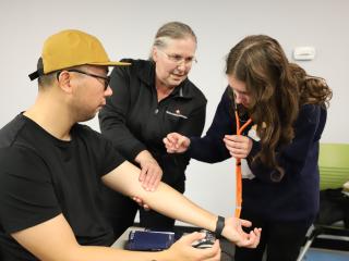 A woman checks a man's pulse, while a teacher watches.