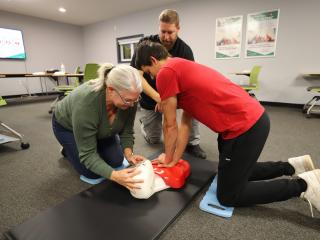 Two students practise CPR on a manikin in a classroom while an instructor guides them.