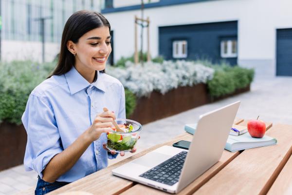 Happy woman holding fresh salad while working on laptop sitting outside office building