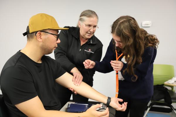 A woman checks a man's pulse, while a teacher watches.