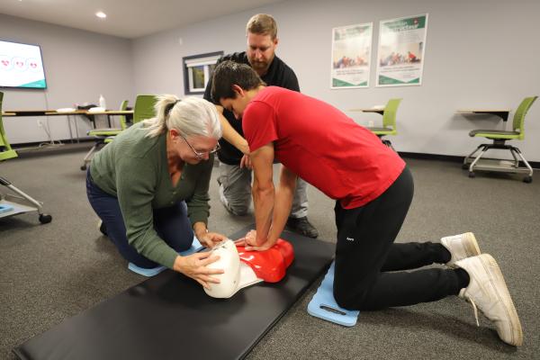 Two students practise CPR on a manikin in a classroom while an instructor guides them.
