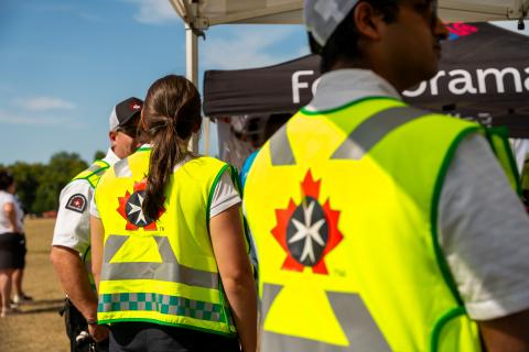 Close-up of St. John Ambulance volunteers wearing yellow safety vests with the organization's eight-pointed cross logo, walking through a festival crowd at Folklorama.