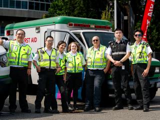 Seven St. John Ambulance first aid volunteers in high-visibility yellow vests and uniforms pose together in front of a white and green St. John Ambulance first aid vehicle at an outdoor event, with a red "First Aid" banner flag behind them.